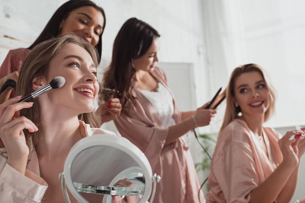 Concentration sélective des femmes multiethniques mettant le maquillage avec une brosse cosmétique et faisant des coiffures dans la chambre à la fête de célibataire
 - Photo, image