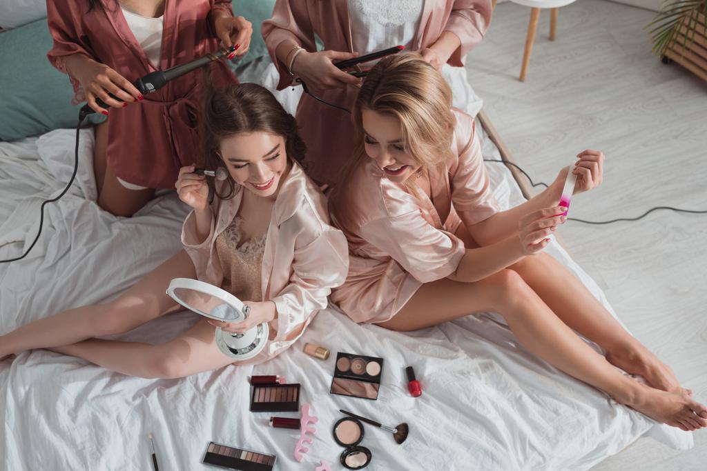 High angle view of multicultural women together using cosmetics and curling irons on bed at bachelorette party - Photo, Image