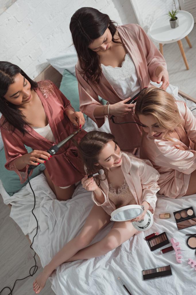 Vista ad alto angolo di donne multietniche insieme utilizzando cosmetici e arricciacapelli sul letto all'addio al nubilato
 - Foto, immagini