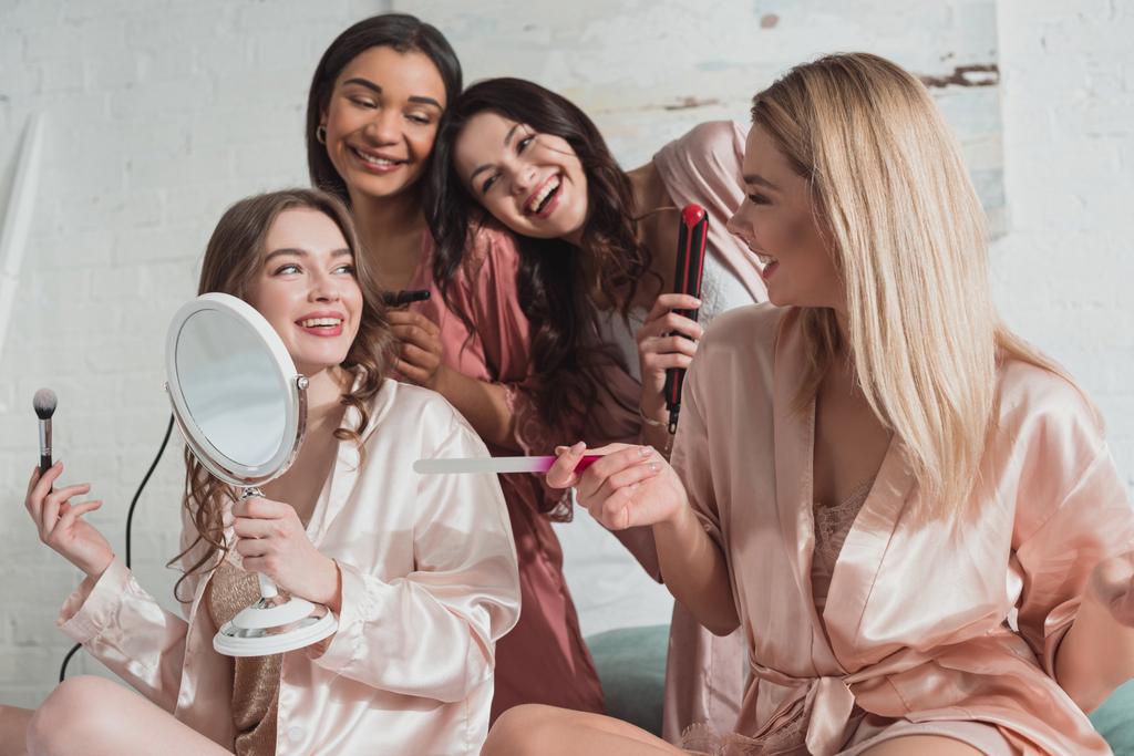 Mujeres multiculturales felices con cepillo cosmético, lima de uñas y rizador mirándose y sonriendo
 - Foto, imagen