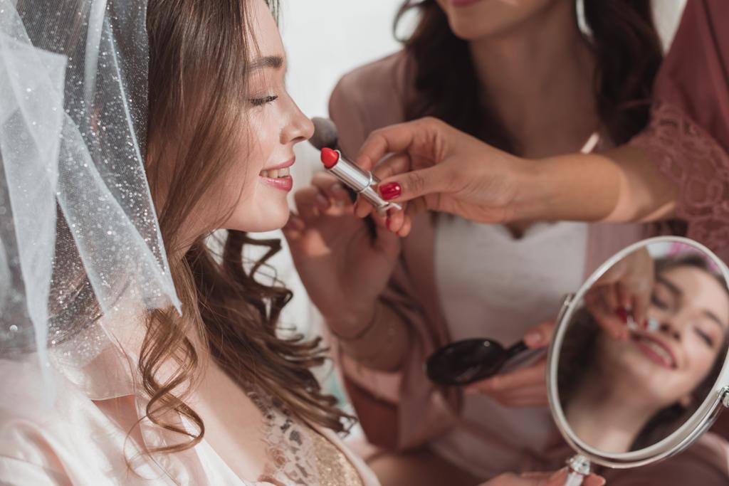 Обрізаний погляд на багатоетнічних жінок, що надягають макіяж косметичним пензлем та помадою на наречену на білому тлі
 - Фото, зображення
