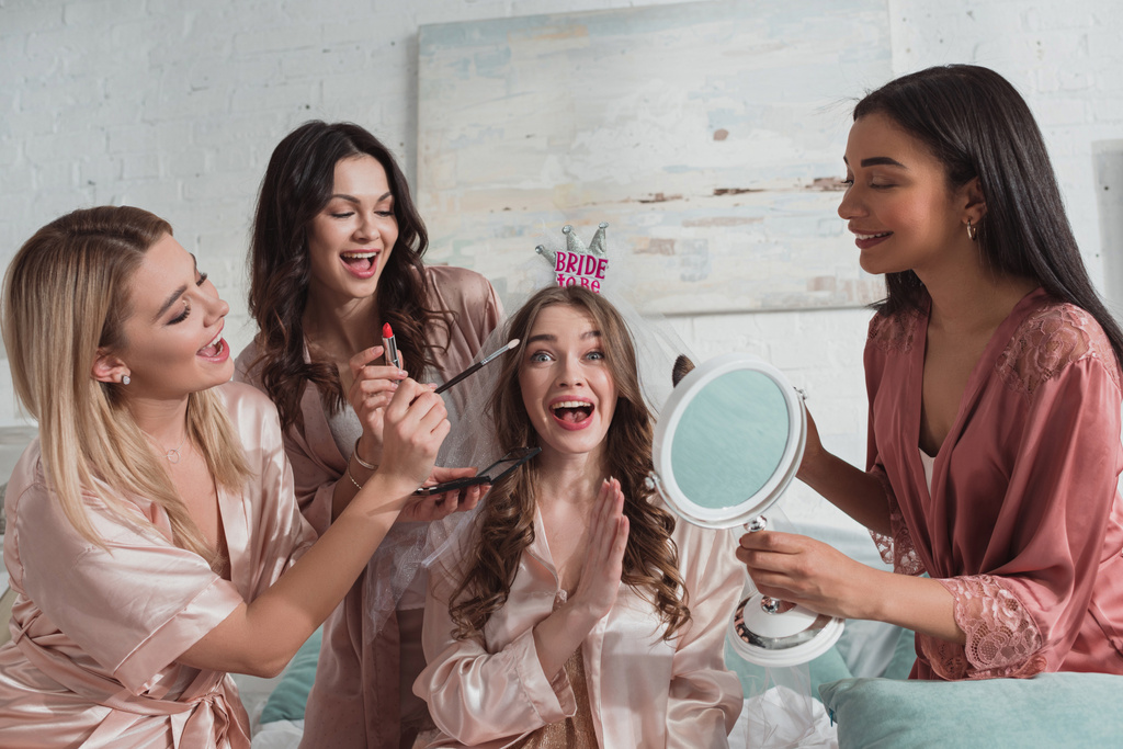 Mujeres multiétnicas que ponen maquillaje con cepillos cosméticos en novia emocionada y feliz con velo nupcial en despedida de soltera
 - Foto, imagen