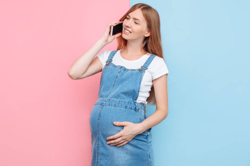 Συναισθηματικά ευτυχισμένη νεαρή έγκυος γυναίκα μιλάει στο τηλέφωνο, στέκεται σε ένα απομονωμένο ροζ και μπλε φόντο - Φωτογραφία, εικόνα