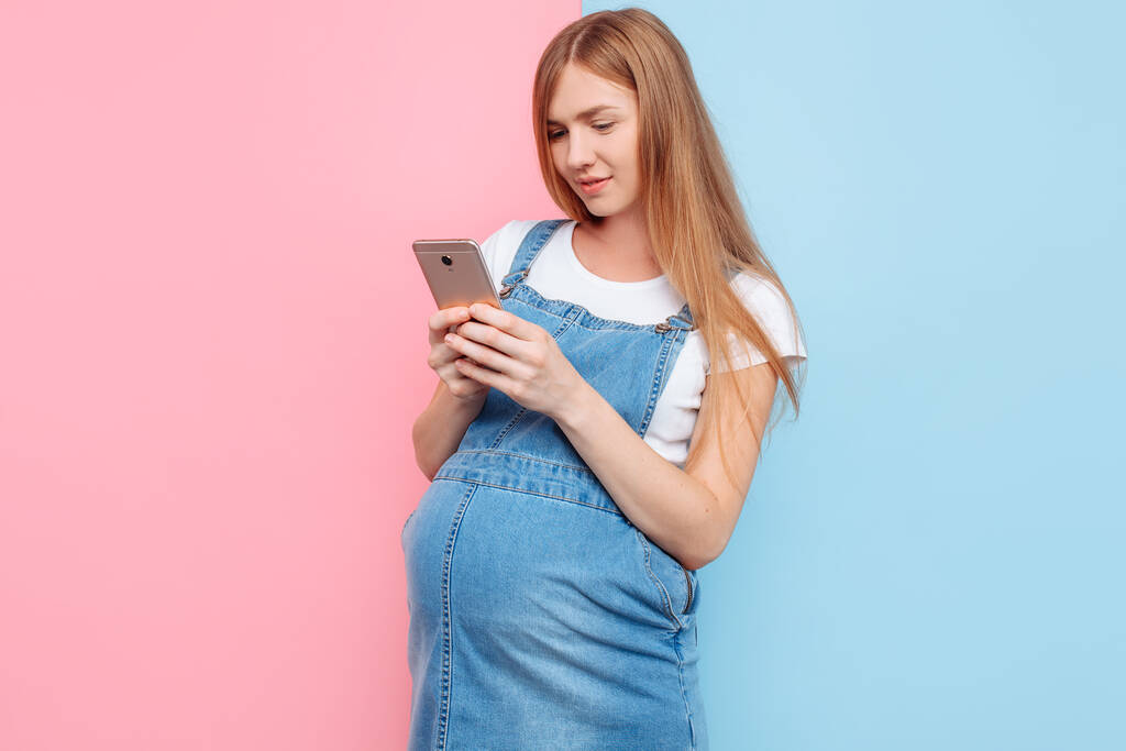 Emotionaalinen onnellinen kaunis nuori raskaana oleva nainen käyttää älypuhelinta seistessään eristetyllä vaaleanpunaisella ja sinisellä taustalla
 - Valokuva, kuva