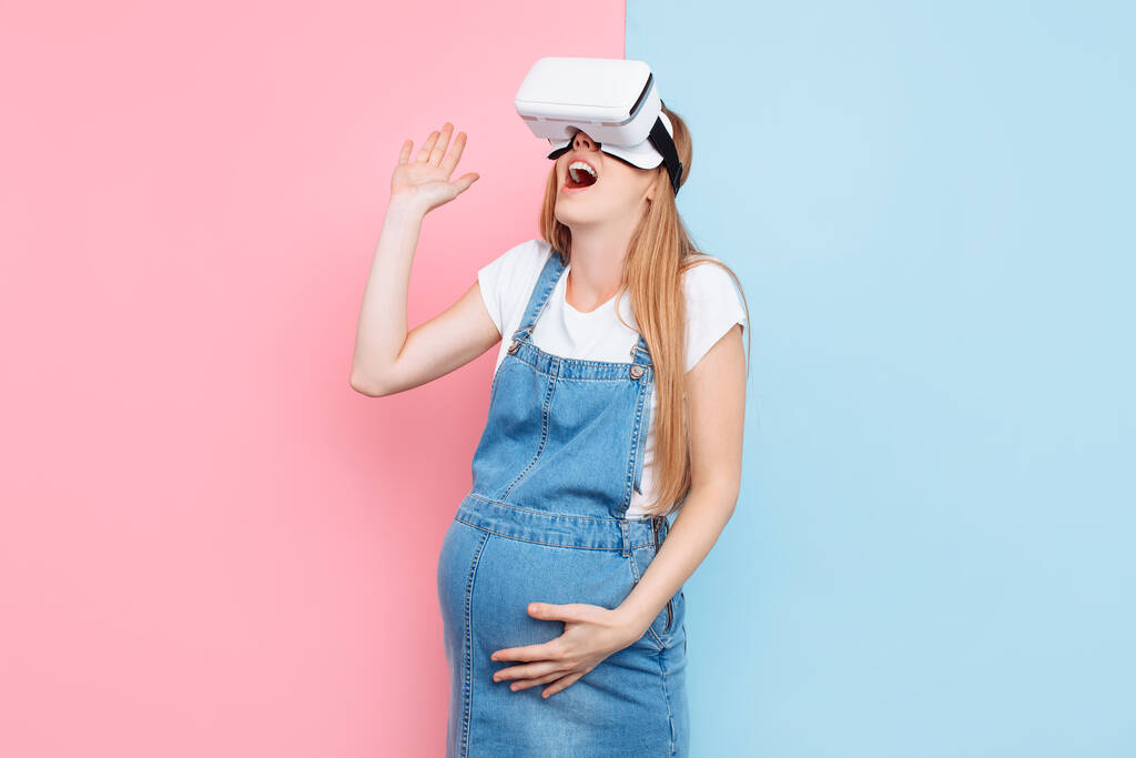 Весела щаслива вагітна жінка весело носить окуляри віртуальної реальності, стоячи на рожевому і синьому фоні
 - Фото, зображення