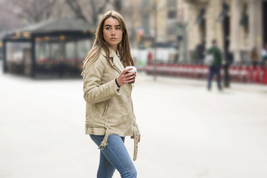 Porträt einer Europäerin, die mit Kaffee zum Mitnehmen in der Hand in einer Straße der Stadt spaziert - Foto, Bild