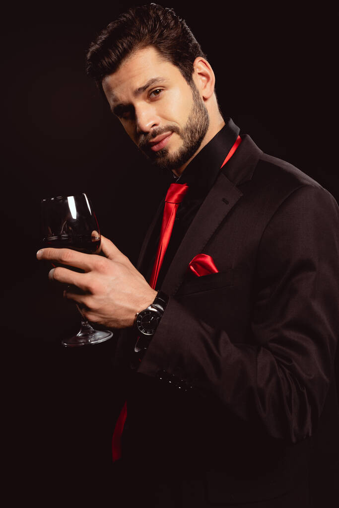 エレガントな若い男は赤ワインのガラスを保持し、黒で隔離されたカメラを見て - 写真・画像