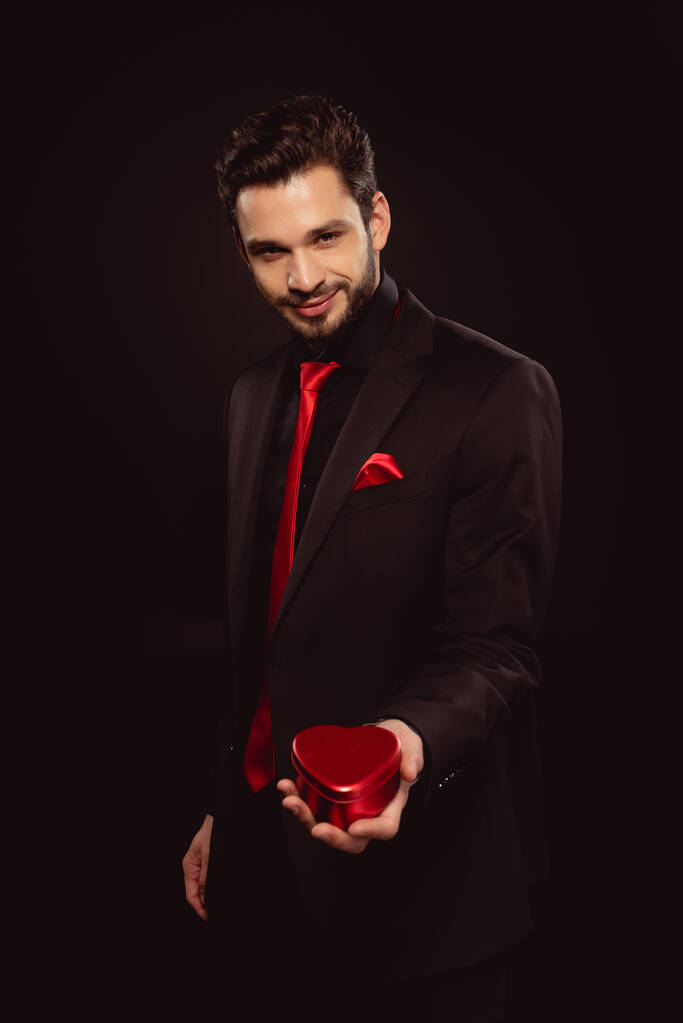 Eleganter Mann im Anzug mit Geschenkbox in Herzform und Blick in die Kamera isoliert auf schwarz - Foto, Bild