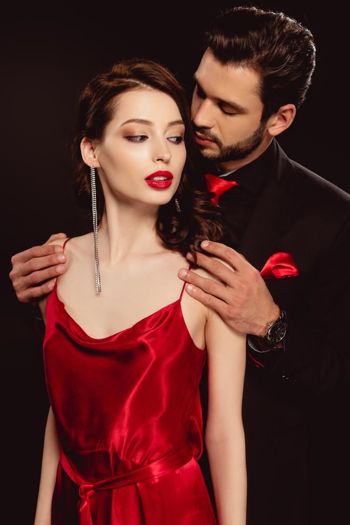 Homme élégant en tenue formelle étreignant belle petite amie en robe rouge isolé sur noir
 - Photo, image