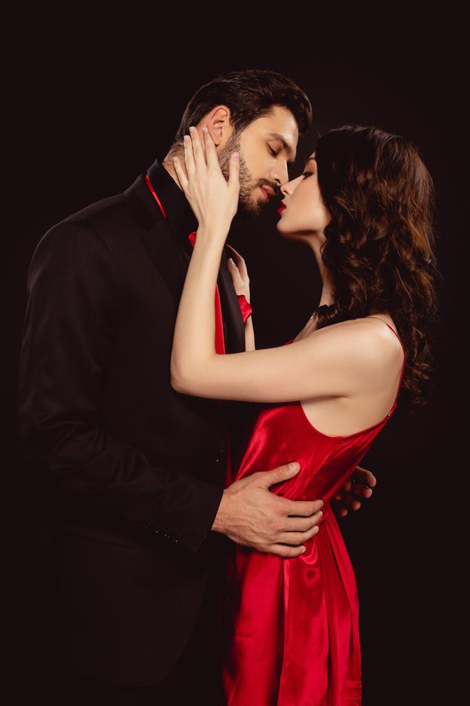 Side view of elegant woman in red dress kissing handsome boyfriend in suit isolated on black - Photo, Image