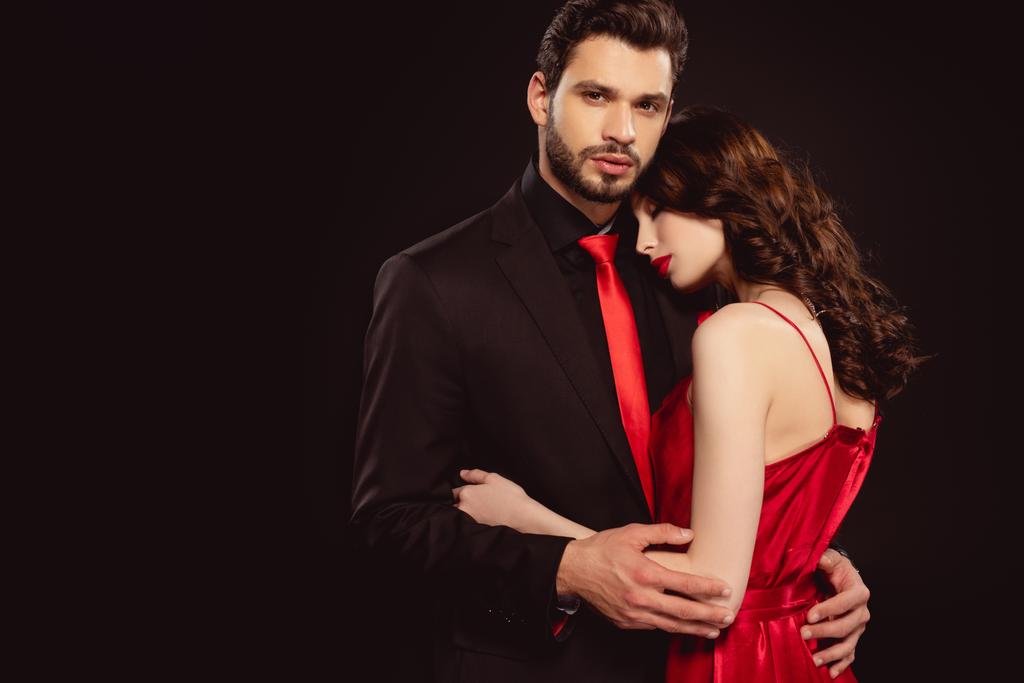 Hombre guapo abrazando a mujer hermosa en vestido rojo y mirando a la cámara aislada en negro
 - Foto, Imagen