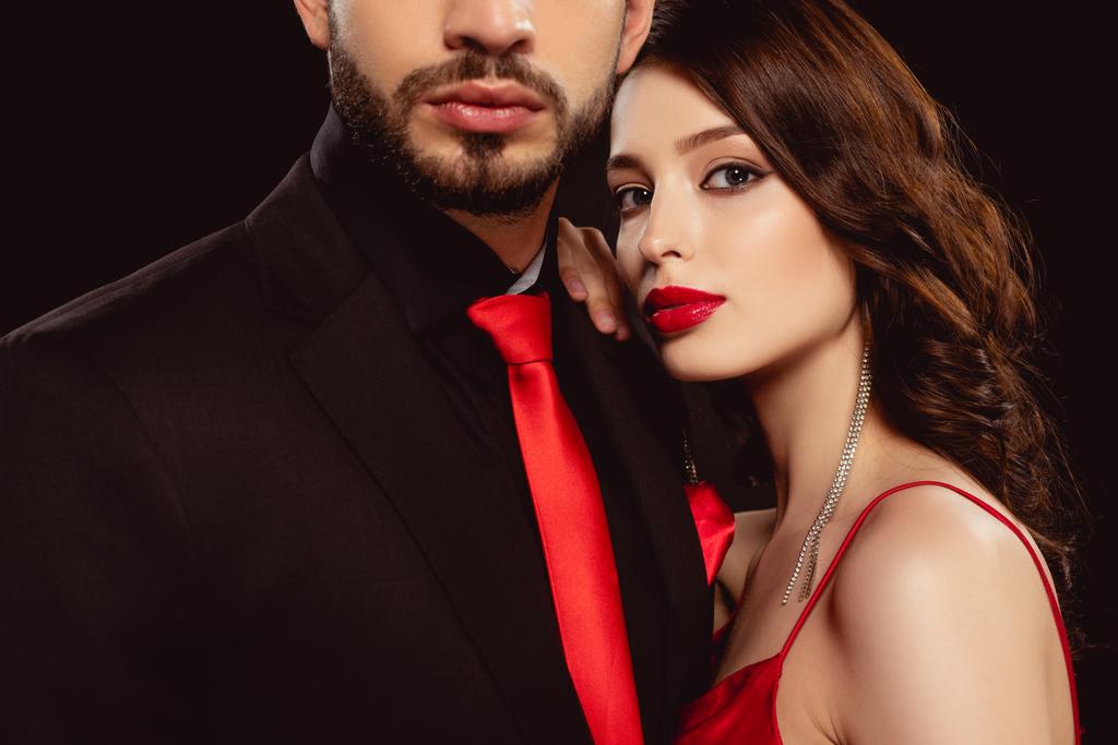 Elegante Frau mit roten Lippen, die in die Kamera schaut, hört Freund in formeller Kleidung isoliert auf schwarz - Foto, Bild