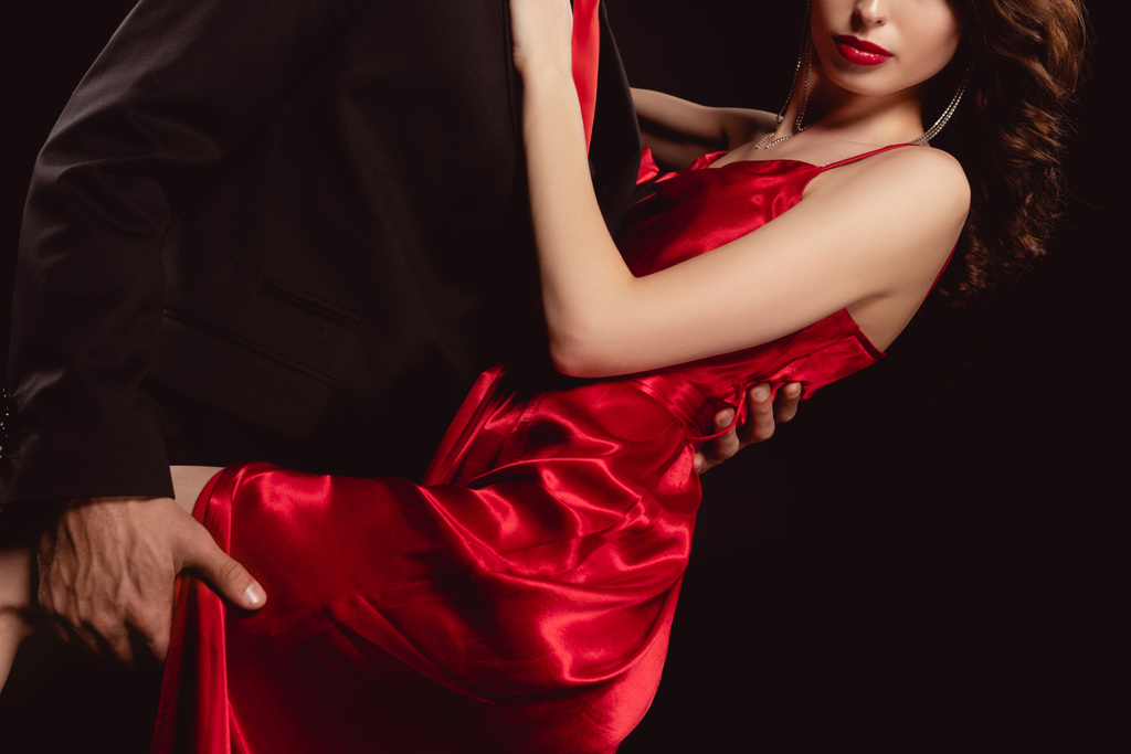 Περικοπή άποψη του κομψού άνδρα αγκαλιάζει και κρατώντας από οδήγησε σαγηνευτική γυναίκα με κόκκινο φόρεμα απομονώνονται σε μαύρο - Φωτογραφία, εικόνα
