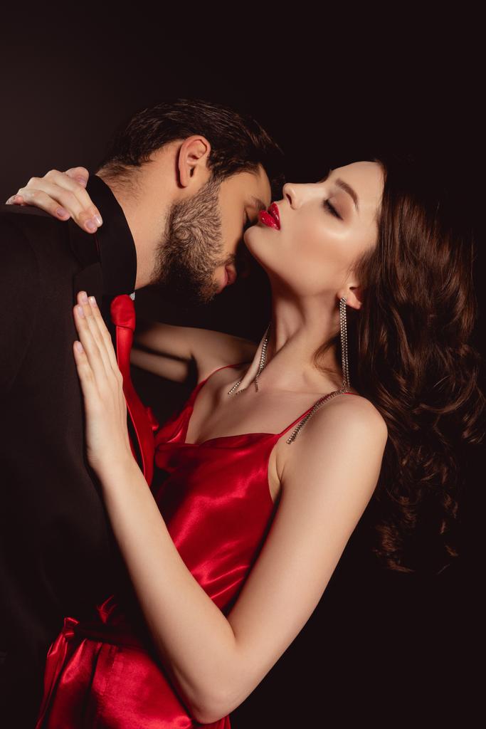 Vista lateral del hombre en traje besar atractiva mujer en vestido rojo aislado en negro
 - Foto, Imagen