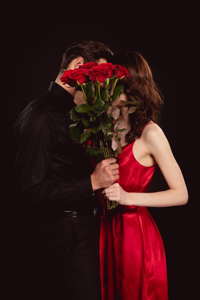 Side view of elegant couple covering faces with bouquet of red roses isolated on black - Photo, Image