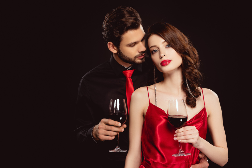 Элегантная женщина с бокалом вина, смотрящая в камеру рядом с красивым парнем, изолированным на черном
 - Фото, изображение