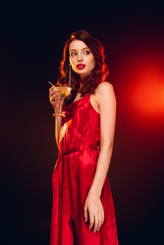 Elegant girl in red dress holding glass of martini on black background with lighting - Photo, Image