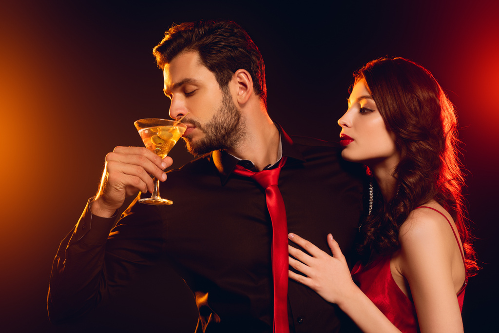 Knappe man drinken cocktail in de buurt elegante vriendin op zwarte achtergrond met verlichting - Foto, afbeelding