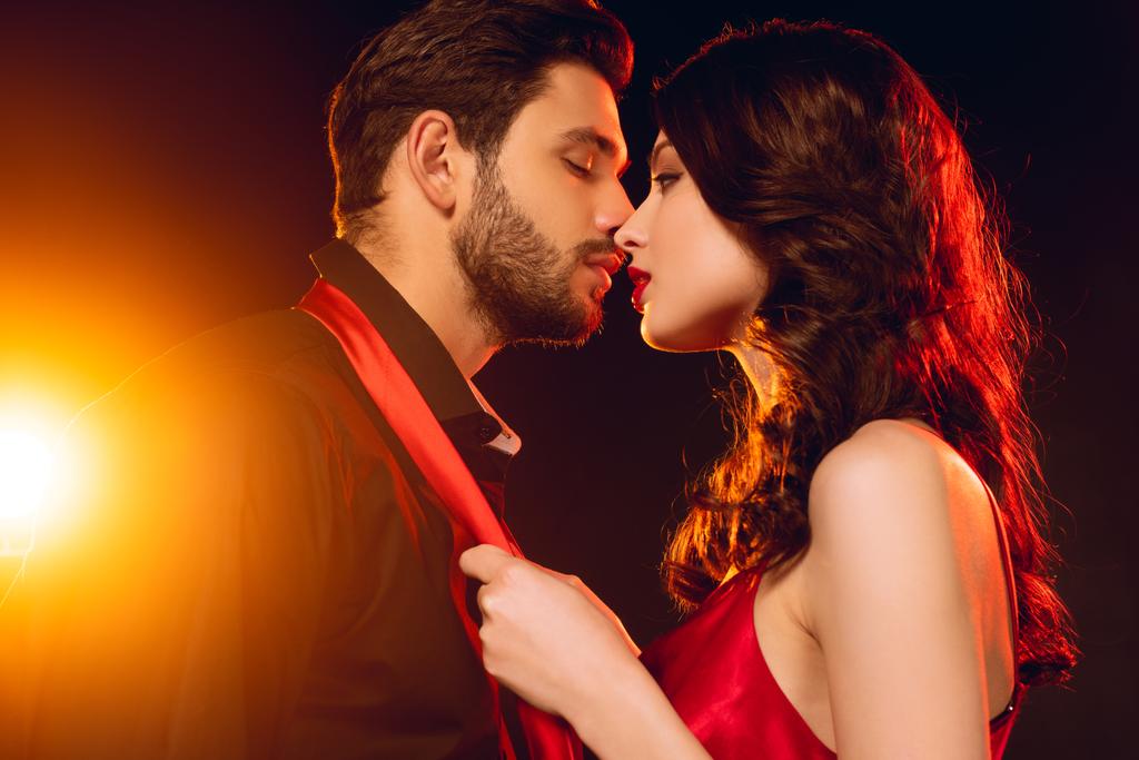 Vue latérale de belle femme embrassant et tenant par cravate beau petit ami sur fond noir avec éclairage
 - Photo, image