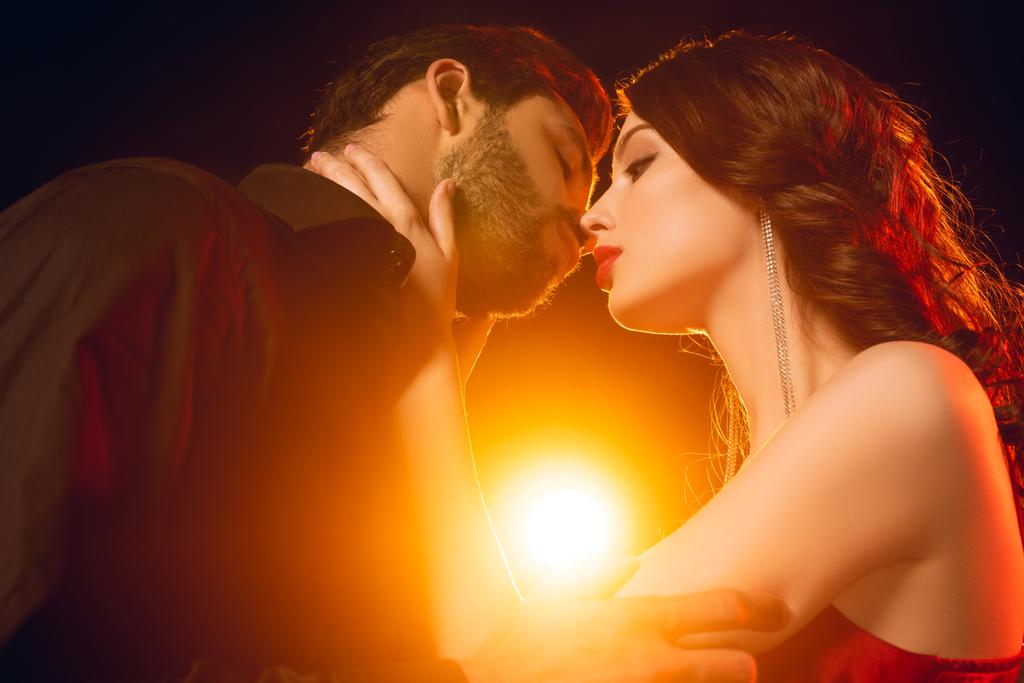 Baixo ângulo vista de mulher elegante beijando bonito namorado no fundo preto com iluminação
 - Foto, Imagem