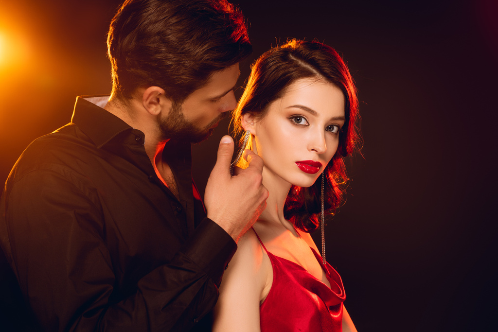 Side view of handsome man touching neck of beautiful girlfriend looking at camera on black background with lighting - Photo, Image