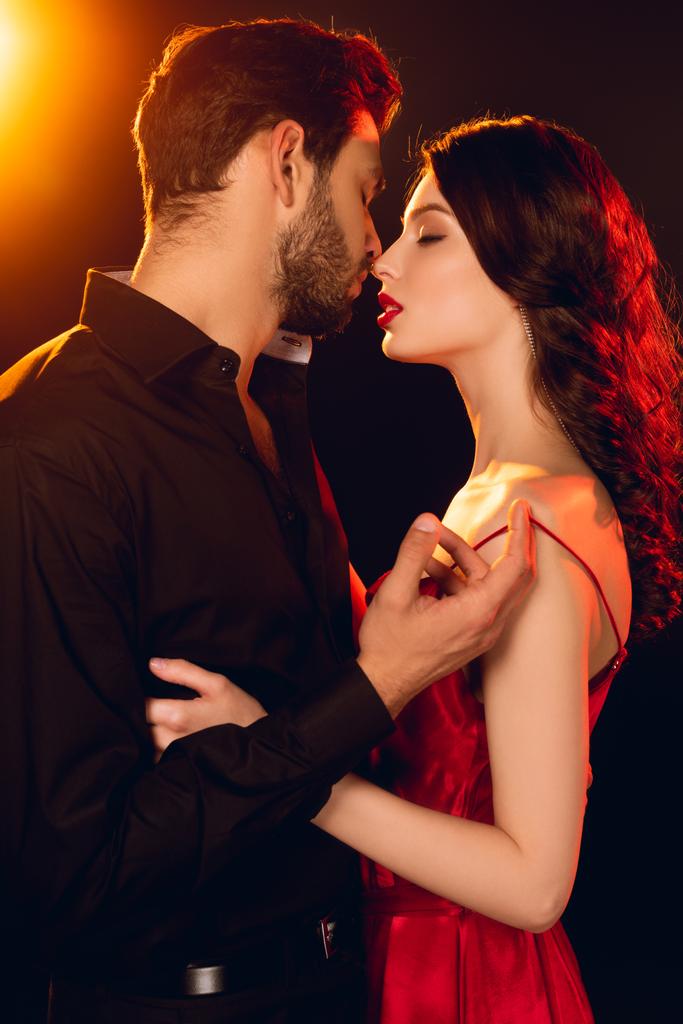 Vista lateral de homem bonito beijando e decolando vestido de bela namorada no fundo preto com iluminação
 - Foto, Imagem