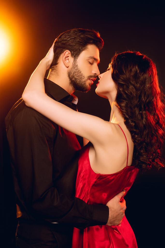 Zijaanzicht van elegante vrouw in rode jurk omarmen en kussen knappe vriend op zwarte achtergrond met verlichting - Foto, afbeelding