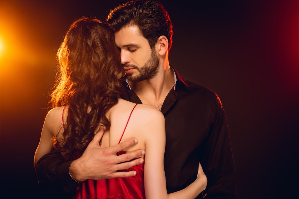 Hombre guapo abrazando novia en vestido rojo sobre fondo negro con iluminación
 - Foto, imagen