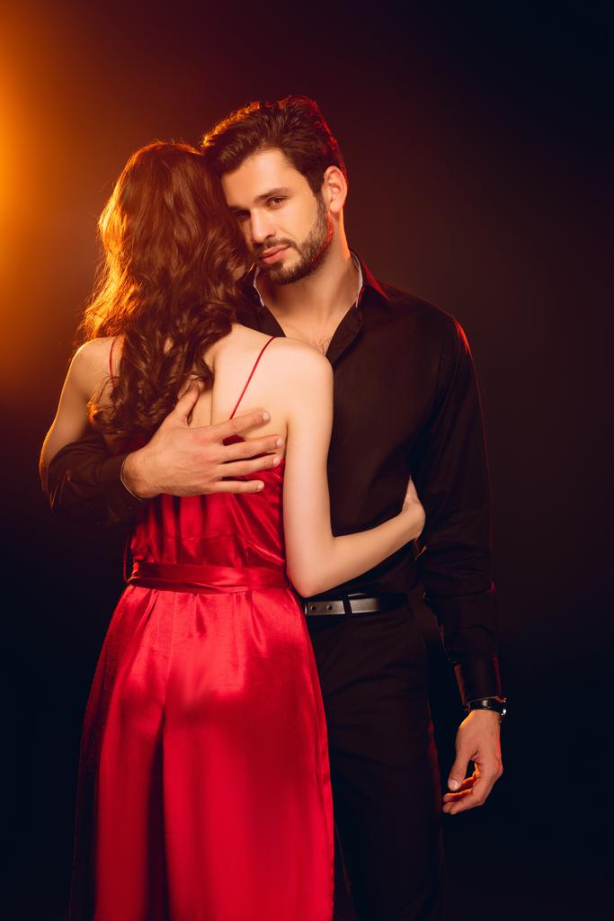 Eleganter Mann blickt in die Kamera, während er Freundin in rotem Kleid auf schwarzem Hintergrund mit Beleuchtung umarmt - Foto, Bild