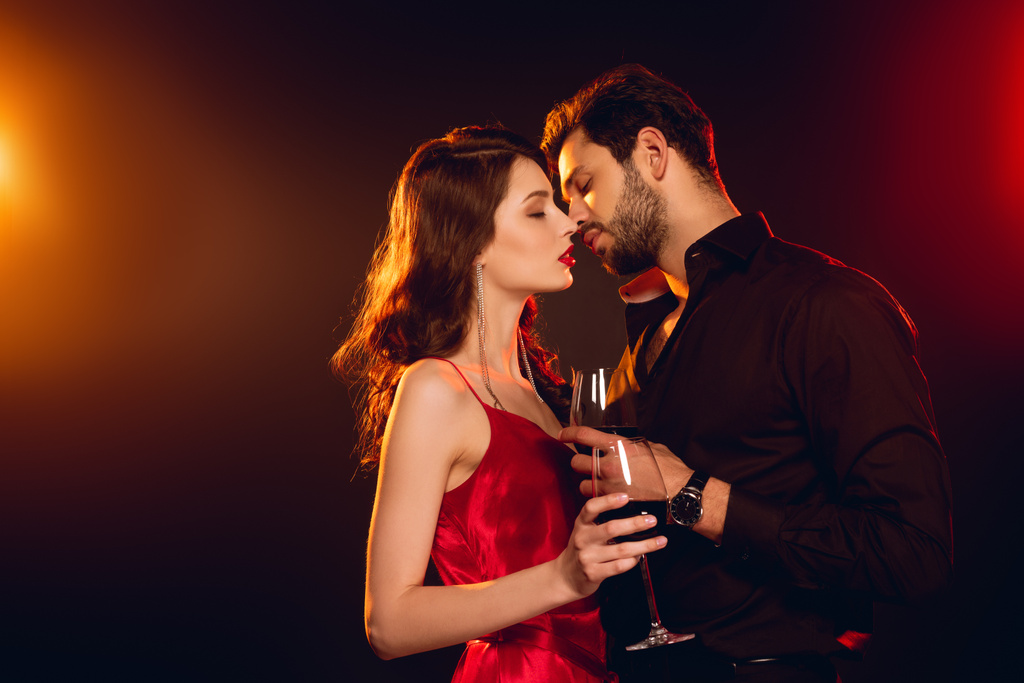 Vista lateral do homem bonito beijando namorada atraente no vestido vermelho com vidro de vinho no fundo preto com iluminação
 - Foto, Imagem