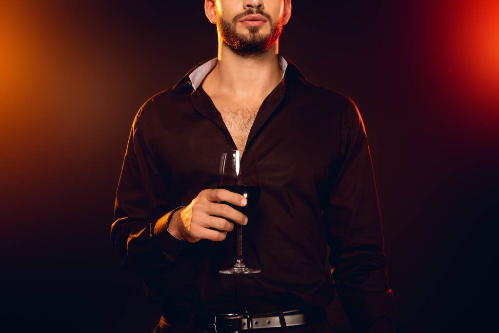 Ausgeschnittene Ansicht eines eleganten Mannes mit einem Glas Rotwein auf schwarzem Hintergrund mit Beleuchtung - Foto, Bild