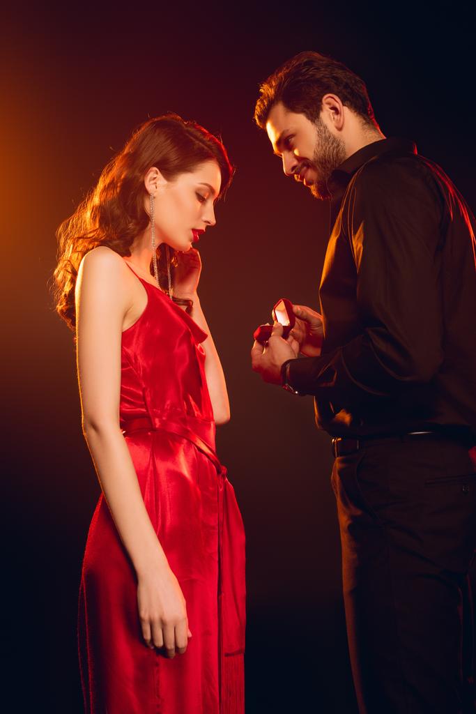 Boční pohled pohledný muž drží dárkové krabice s šperky prsten v blízkosti krásné přítelkyně na černém pozadí s osvětlením - Fotografie, Obrázek