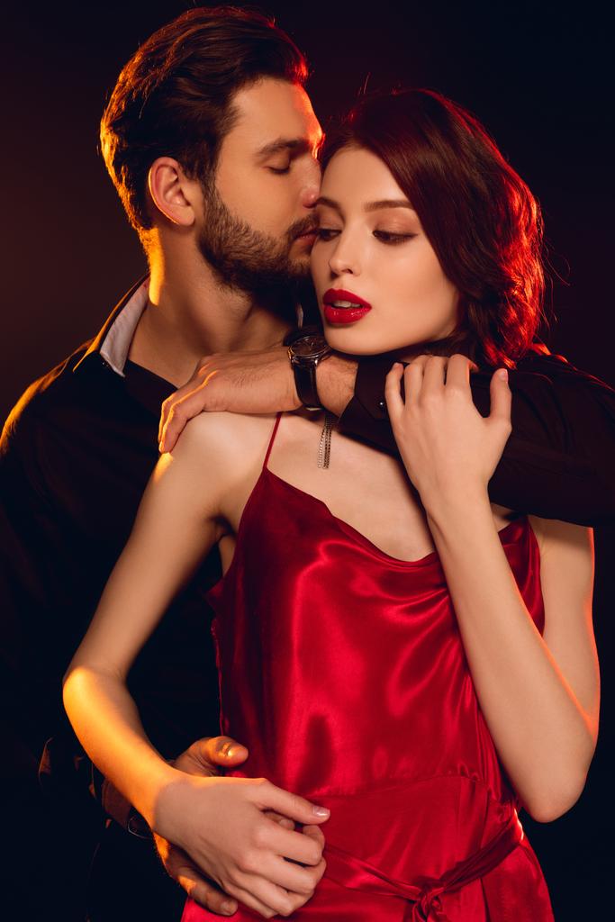 Hombre guapo abrazando y besando novia elegante en vestido rojo aislado en negro
 - Foto, Imagen