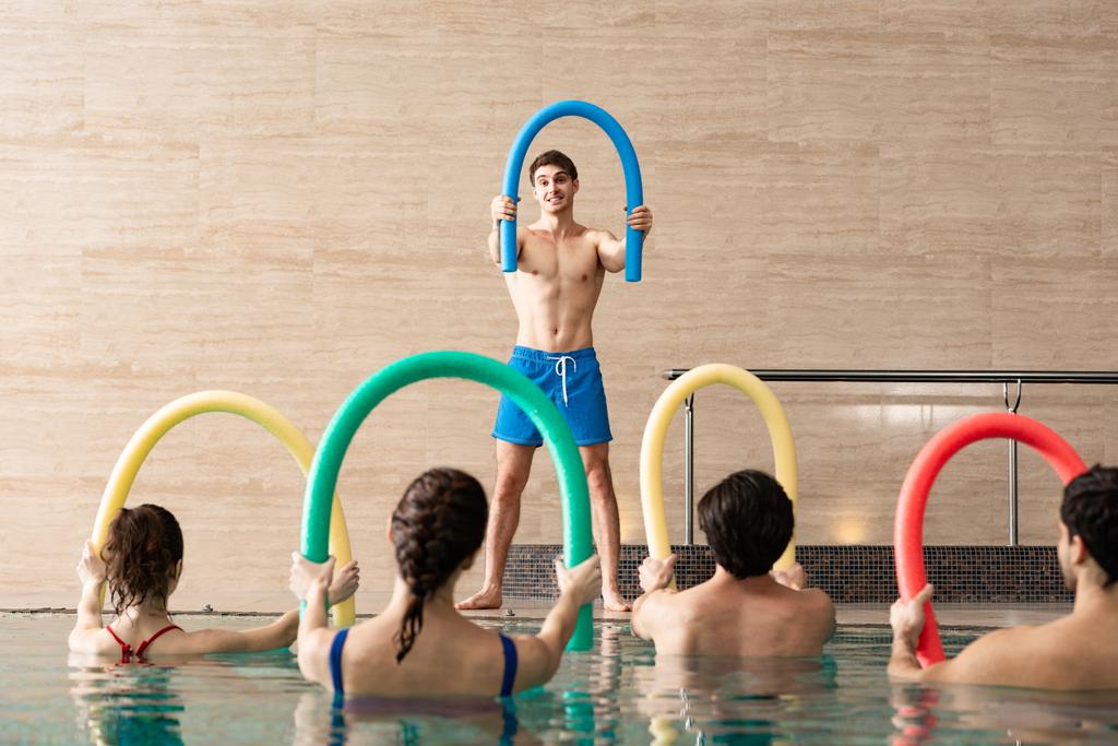 Grupo de jovens que se exercitam com macarrão de piscina e bonito treinador na piscina
 - Foto, Imagem