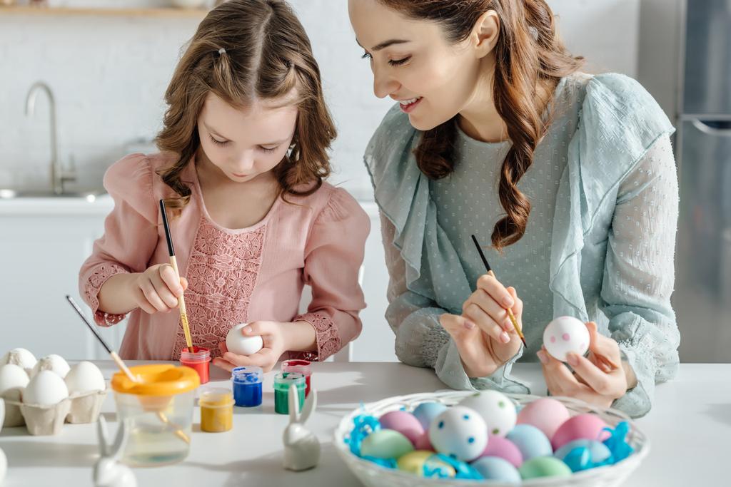 selective focus of happy mother and daughter painting easter eggs near decorative rabbits  - Photo, Image