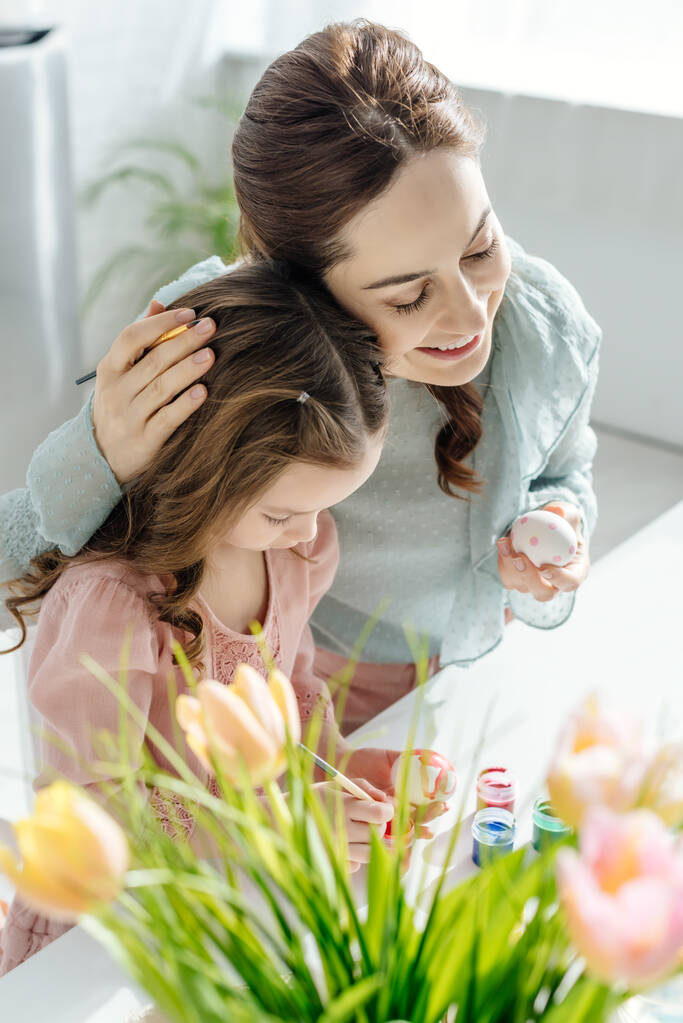 Selektiver Fokus der glücklichen Mutter, die ihre süße Tochter umarmt und neben Tulpen Ostereier bemalt  - Foto, Bild