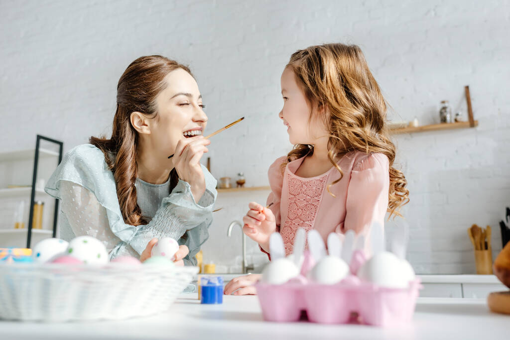 επιλεκτική εστίαση χαρούμενη μητέρα και ευτυχισμένη κόρη κοντά ζωγραφισμένα πασχαλινά αυγά  - Φωτογραφία, εικόνα