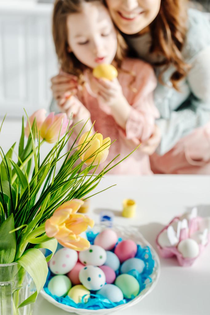Mutlu bir anne ve Paskalya yumurtası boyayan tatlı bir kızın yanındaki lalelerin seçici odağı.  - Fotoğraf, Görsel