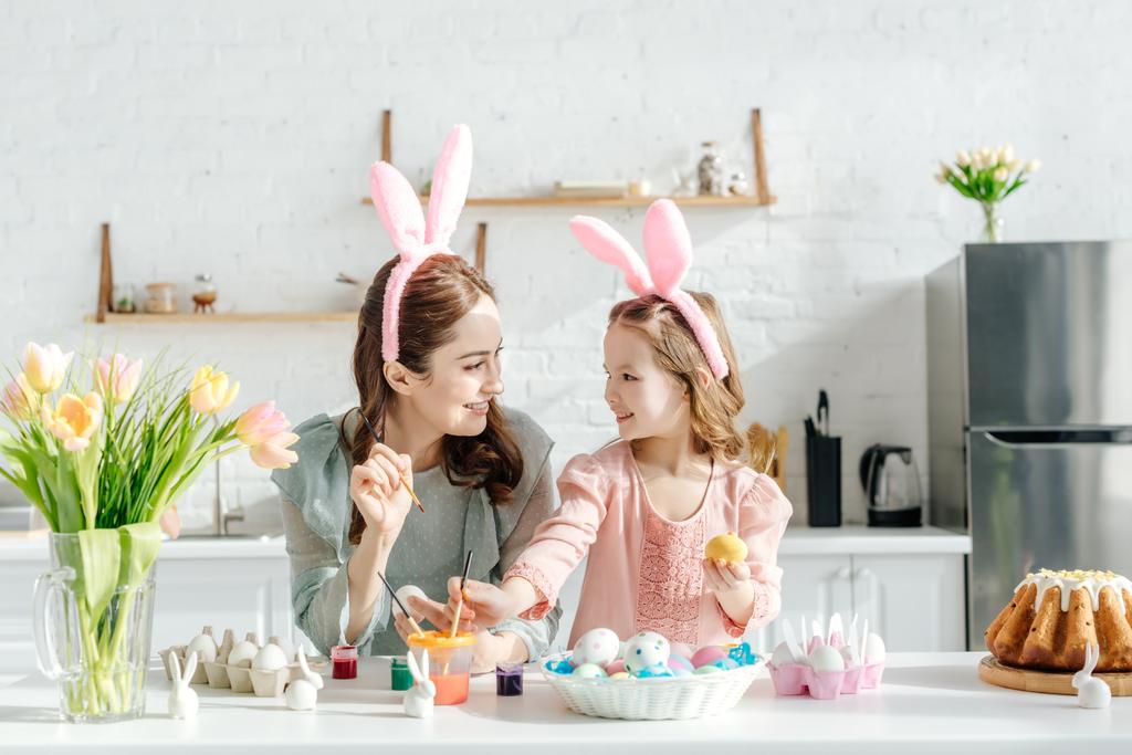 happy child and mother with bunny ears near chicken eggs, decorative rabbits, easter bread and tulips  - Photo, Image
