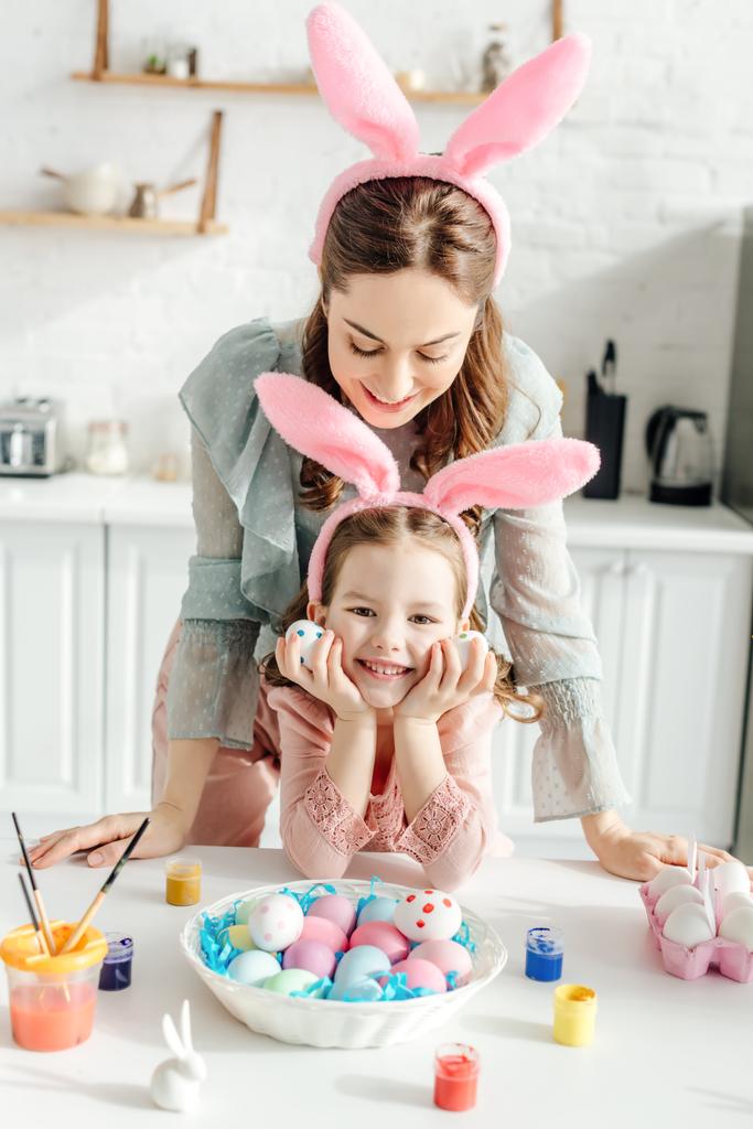 šťastná matka při pohledu na králičí uši na dceru v blízkosti velikonočních vajec  - Fotografie, Obrázek