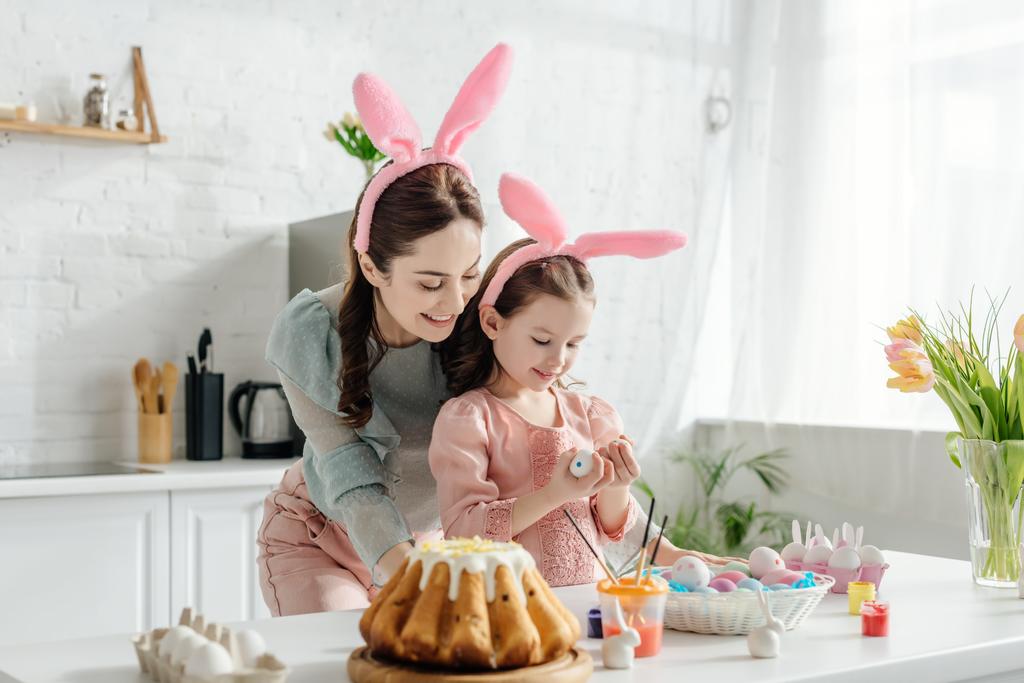 Ευτυχισμένη μητέρα και κόρη στα αυτιά κουνελιού κοιτάζοντας αυγά κότας κοντά σε τουλίπες και πασχαλινό ψωμί  - Φωτογραφία, εικόνα