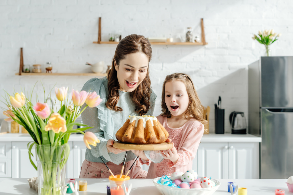 ενθουσιασμένοι μητέρα και παιδί κοιτάζοντας ψωμί Πάσχα κοντά σε αυγά Πάσχα, διακοσμητικά κουνέλια και τουλίπες  - Φωτογραφία, εικόνα
