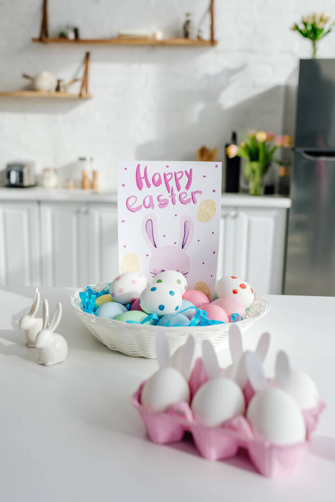 foco seletivo de ovos de galinha pintados perto de coelhos decorativos e cartão de saudação com letras felizes da Páscoa
  - Foto, Imagem