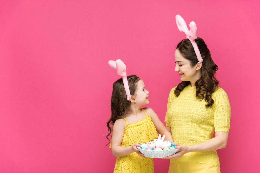 happy mother and daughter in bunny ears holding easter eggs and looking at each other isolated on pink  - Photo, Image