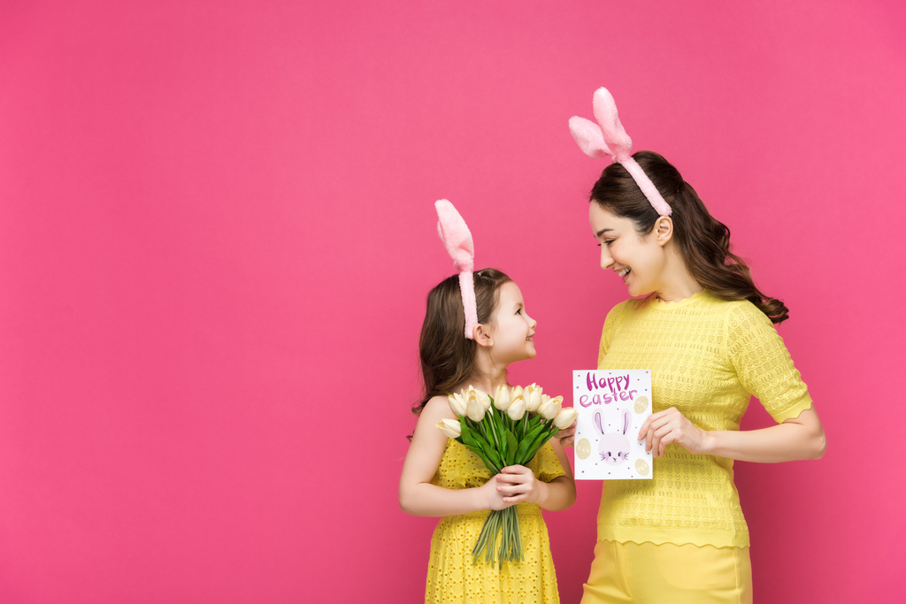 mãe alegre em orelhas de coelho segurando cartão de saudação com feliz Páscoa lettering perto da filha com tulipas isoladas em rosa
  - Foto, Imagem