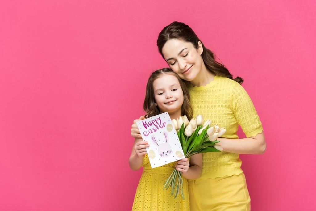 vrolijke moeder met wenskaart met vrolijke paaslettering en knuffelende dochter met tulpen geïsoleerd op roze  - Foto, afbeelding