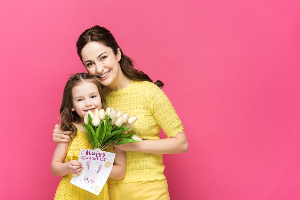 veselá matka drží blahopřání se šťastnými velikonočními písmeny a objímající dítě s tulipány izolované na růžové  - Fotografie, Obrázek
