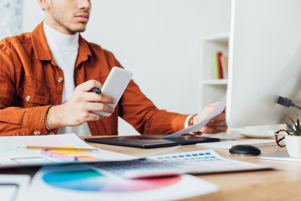 Обрезанный вид ux дизайнер держа смартфон во время работы с веб-сайта эскизы и цветовой палитры за столом в офисе
  - Фото, изображение