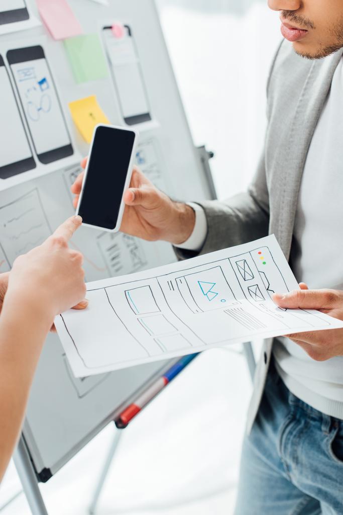 Обрезанный вид дизайнера указывающий на смартфон рядом с коллегой с шаблонами ux дизайна возле доски в офисе
  - Фото, изображение