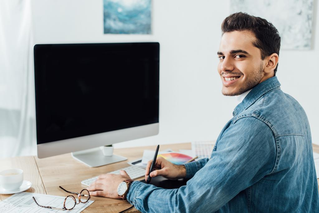 Dizájner mosolyog a kamera, miközben dolgozik a projekt felhasználói élmény tervezés közelében számítógép és grafika táblagép az asztalon  - Fotó, kép
